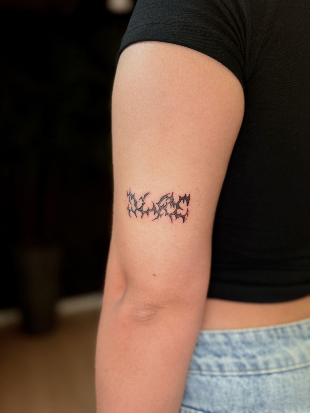 Palavra em lettering de traço tribal com pontas agudas e preenchimento preto, na parte de trás do braço. Por Dudis (Maria Eduarda Freitas), na ADV Tattoo.
