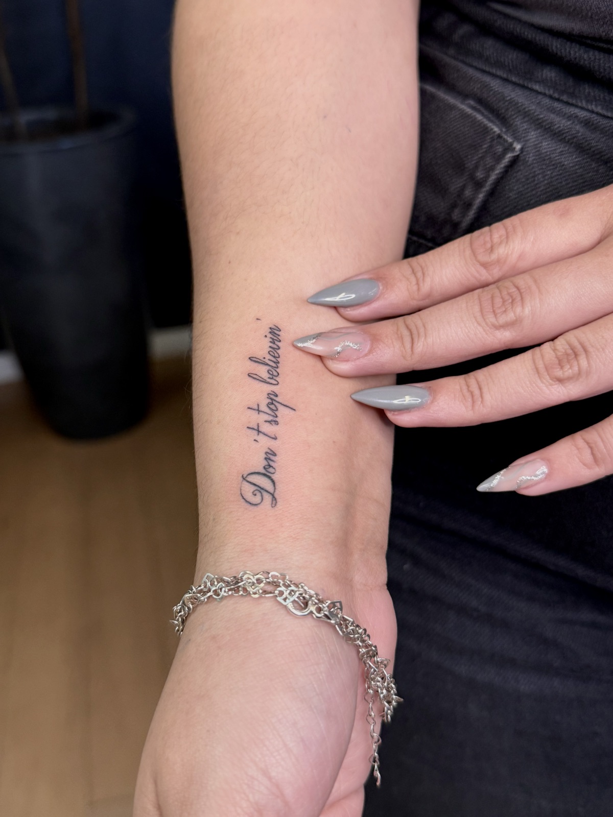 Frase "Don’t stop believin’" em letra cursiva de traço fino, no antebraço perto do punho. Por Dudis (Maria Eduarda Freitas), na ADV Tattoo.